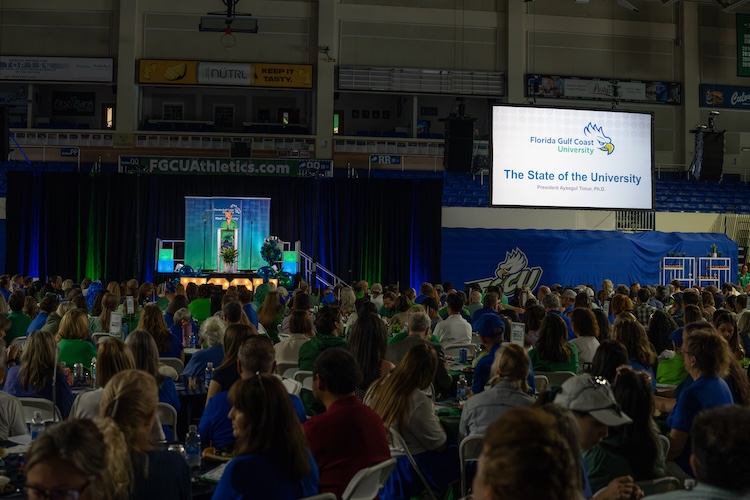 A large audience sits at tables inside Alico Arena as they watch President Timur gives remarks. A screen to the right side next of the stage displays “The State of the University” alongside the Florida Gulf Coast University logo. President Timur is seen on stage. She is wearing a green blazer and blue skirt.