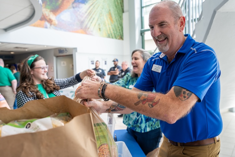 Dressed in a blue polo, Mike Stavely, is standing behind a table collecting food donations for the FGCU Campus Food Pantry. Other employees can be seen in the back handing over their donations.