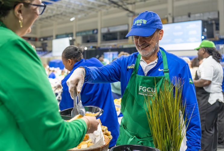 Vice Provost Paul Dosal is using tongs to place hot food on a plate behind held by an employee who is wearing a green jacket. He is wearing a blue FGCU hat, a white shirt, with a blue FGCU branded sweater and a green apron with the words Dining Services in white on top.