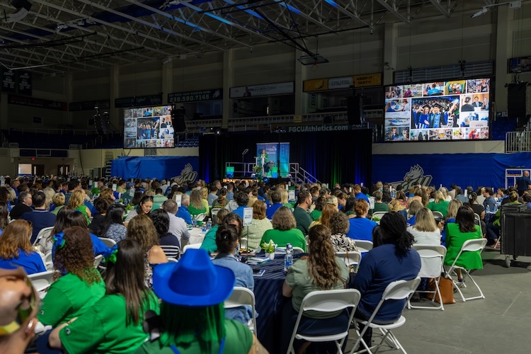 Panoramic view of FGCU employees at Alico Arena dressed in green and blue sitting down at their tables as they listen to President Timur give remarks. Large screens can be seen on the left and ride side of the arena displaying a collage of employee photos.