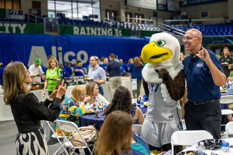 An FGCU employee dressed in a black and white dress with a black sweater on top is taking a photo of the FGCU mascot Azul with an FGCU employee, who is wearing a dark blue polo and black pants. People walking by with plates of food as well as sitting down at tables can be seen in the background.