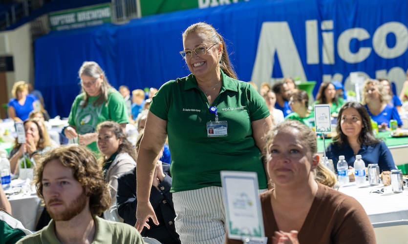 An FGCU employee dressed in a green polo and white pants is standing up among the crowd of people sitting down at their tables inside Alico Area. Two other employees can be seen in the background standing up as well.