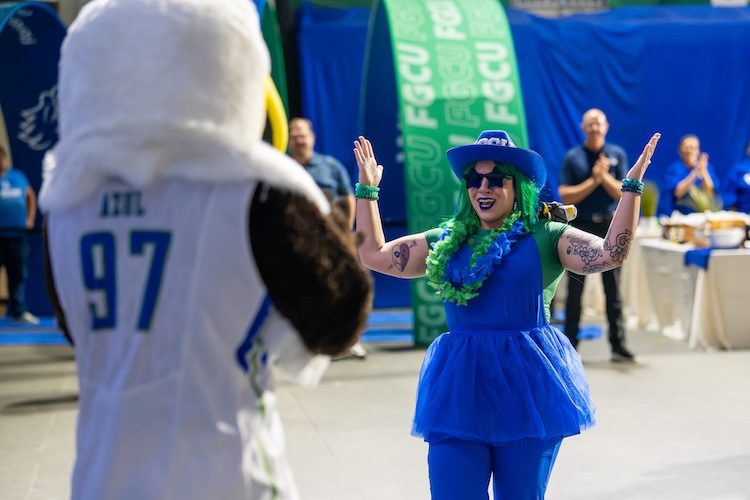 An FGCU employee dressed in blue and green clothing, including a blue hat and green lei, stands with both hands raised as she walks toward FGCU Eagle mascot Azul, who is wearing a white jersey displaying the number 97. Several attendees stand in the background near tables and green banners with the letters FGCU in white.