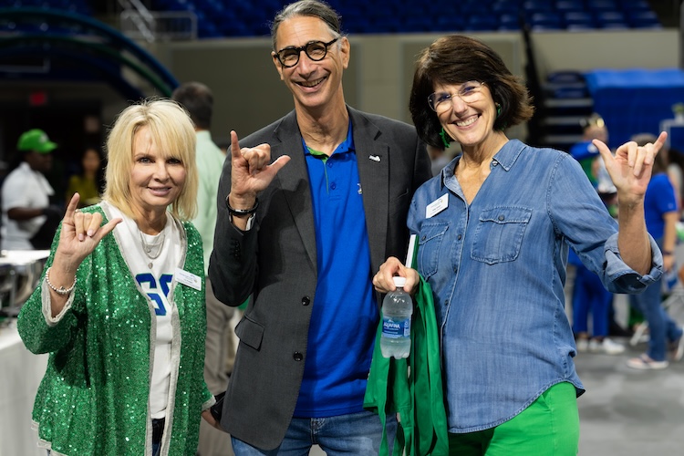 Three FGCU employees stand side by side inside Alico Arena at the Fall Kickoff event, facing the camera and making the “Wings Up” salute. The employee on the left wears a green sequined cardigan over a white FGCU shirt, the person in the middle wears a blue polo shirt and dark blazer, and the person on the right wears a denim shirt while holding a water bottle. Additional attendees are visible in the background.