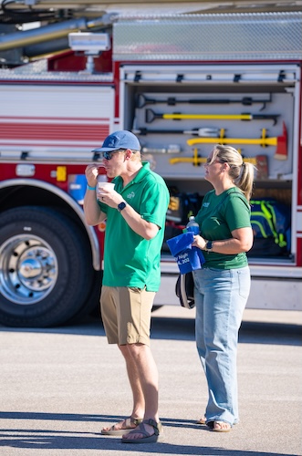 Two people stand beside a fire truck with an open equipment compartment. One person is eating an ice cream while the other holds a blue bag.Both are wearing green shirts.