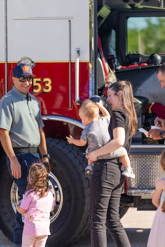 A family stands next to a fire truck and a firefighter, who is showing them the truck. One adult holds a small child while another child stands nearby, and the truck’s open door reveals equipment inside.