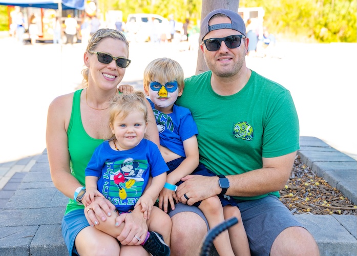 A family of four sits closely together on a stone wall outside Swanson Stadium, dressed in green and blue. One child has their face painted as an eagle.