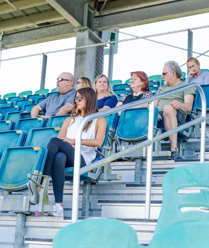FGCU employees and spectators are sitting down at the Swanson Stadium bleachers as they watch a baseball game.