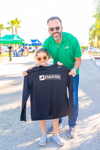GCU employee dressed in a green polo shirt poses for a photo with his child as she holds up a dark blue long-sleeved athletics shirt.