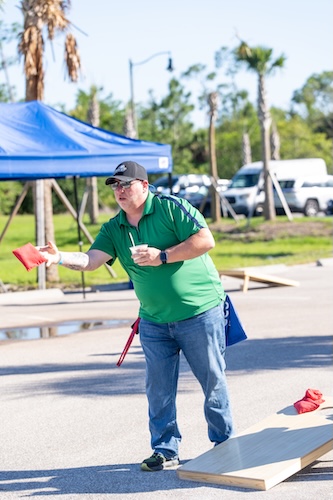FGCU employee, wearing a black hat, and a green and blue polo shirt, is playing corn hole outside of Swanson Stadium.