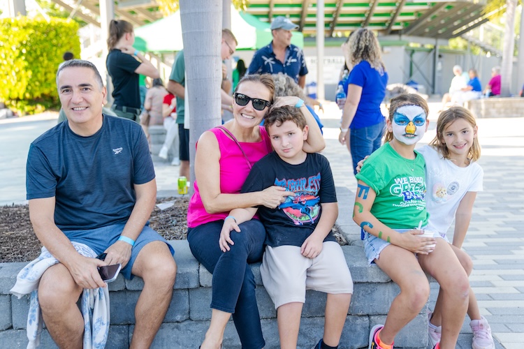 A family of five sit outside at Swanson Stadium. One person wears FGCU Eagle face paint, and others wear casual summer clothing. A pavilion and several people are visible in the background.