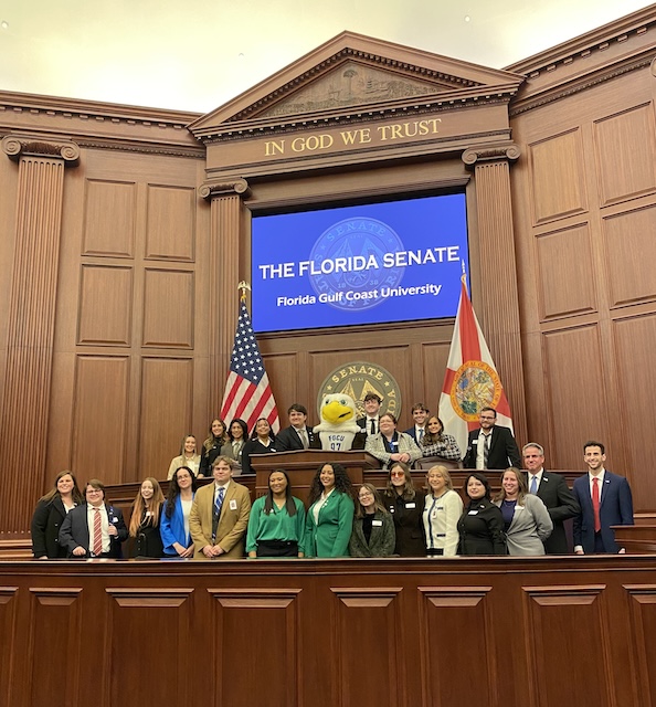 A large group of FGCU students stands together at the front of the Florida Senate chamber beneath the “In God We Trust” inscription. The group is positioned in front of the dais, with U.S. and Florida flags displayed behind them and a screen reading “The Florida Senate – Florida Gulf Coast University.” They are dressed in business attire.  