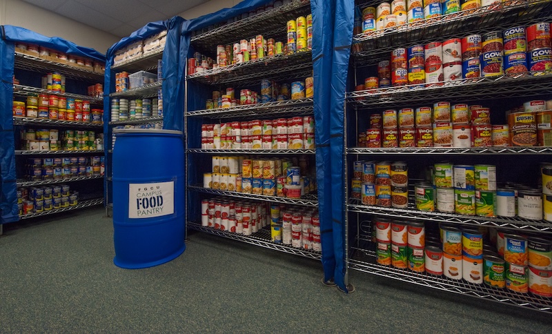 View of the inside the FGCU Campus Food Pantry. The image shows a room with multiple shelves stocked with canned goods and a blue barrel with the words “Campus Food Pantry” on it. 