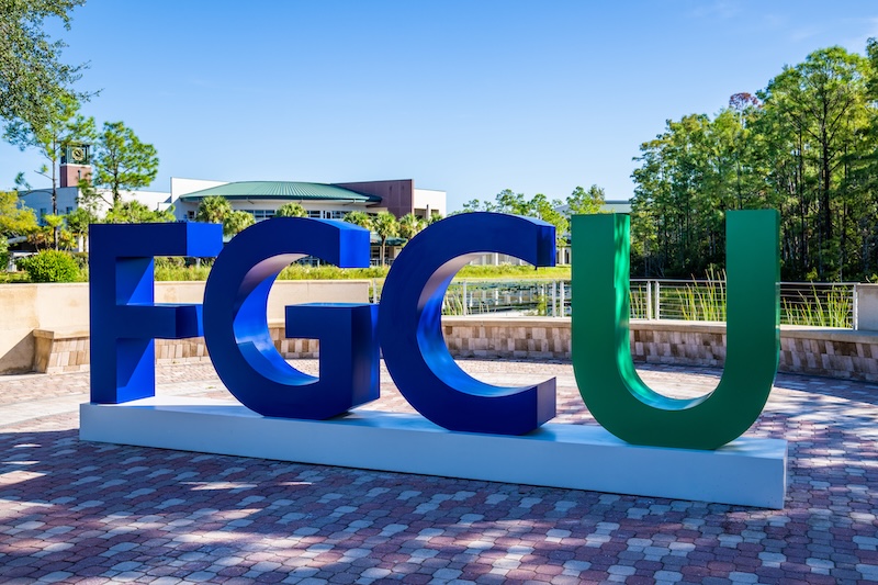 Large, three-dimensional letters spelling "FGCU" in blue and green colors. These letters are mounted on a white base and situated outdoors on a paved area at the Library Lawn. In the background, there are trees, greenery, and a partial view of the Cohen Student Union. 