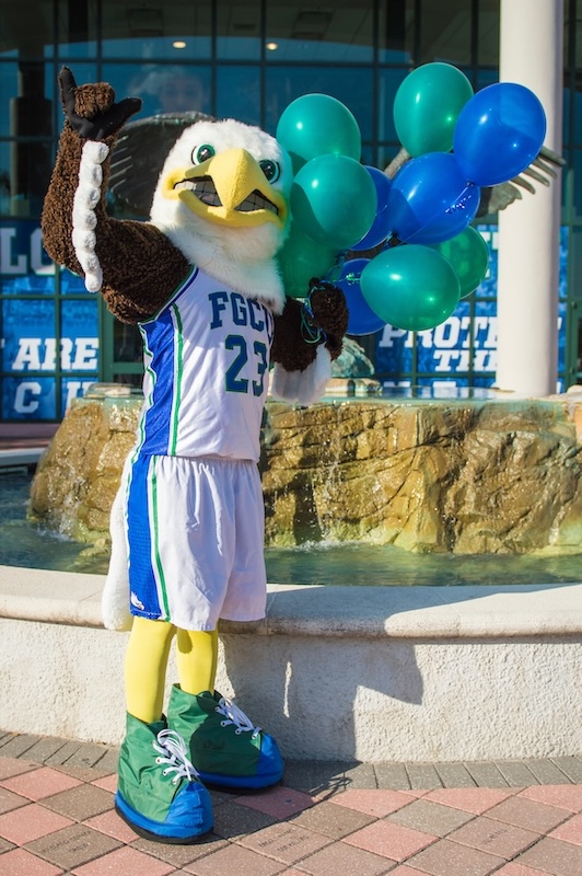 Standing behind Alico arena and its water fountain is FGCU’s mascot Azul. He is holding green and blue balloons with his left hand and is doing the Wings Up salute with his right hand. Azul is wearing a white jersey uniform with blue and green stripes on the sides and the letters FGCU and number 23 on the front of the jersey. 