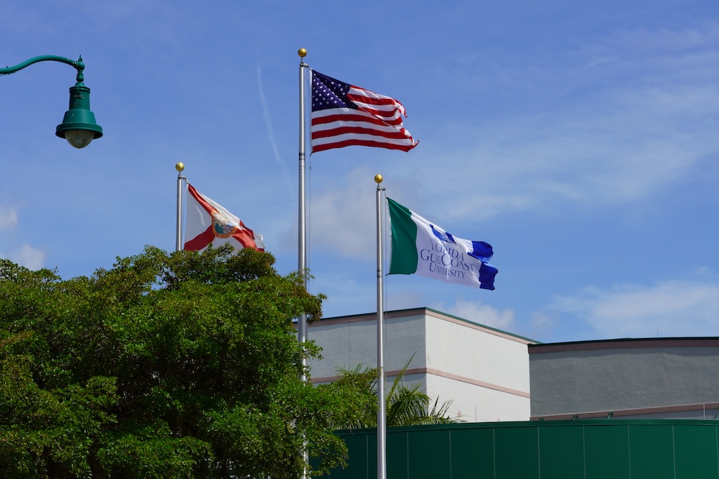 A close-up of the state of Florida, United States and FGCU flags that fly outside Alico Arena on a sunny day. Trees can be seen in the background.  