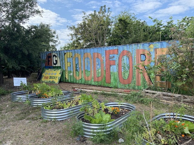 A garden features five raised metal beds filled with plants and flowers. A colorful mural on the wall of a container is in the background reading 'FOOD FOREST.’ The garden is surrounded by trees and greenery under a partly cloudy sky. 