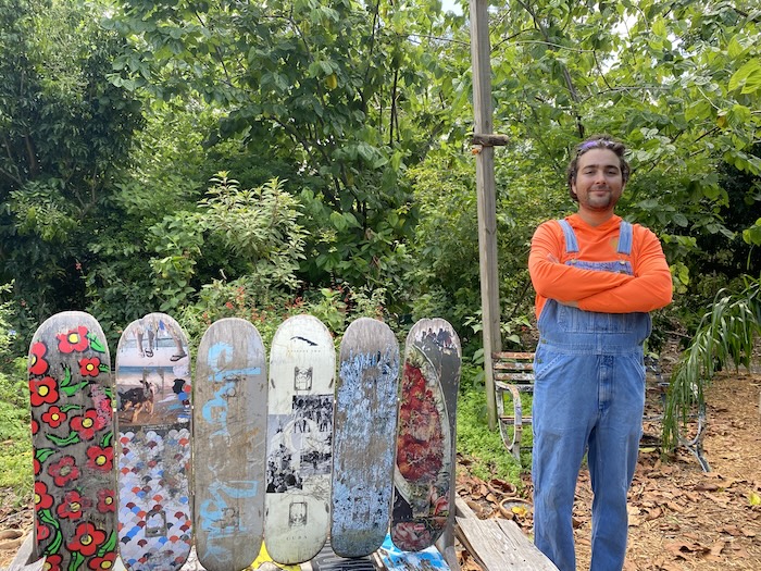 Wearing a bright orange long sleeve shirt and jean overalls, Food Forest manager Dylan Moncada is standing inside the Food Forrest next to a bench made from wooden boards painted with various colors and patterns. 