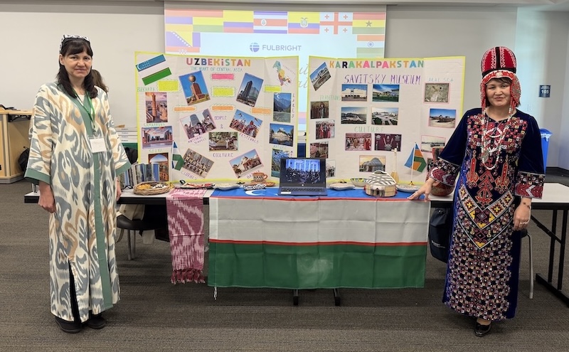 Two individuals from the Fullbright Teaching Excellence Achievement Program are wearing traditional garments and stand beside a cultural display table featuring posters titled “Uzbekistan” and “Karakalpakstan.” The table is decorated with regional textiles, flags, decorative plates, and a laptop showing additional imagery. Colorful photos and maps are arranged on the posters behind them.  