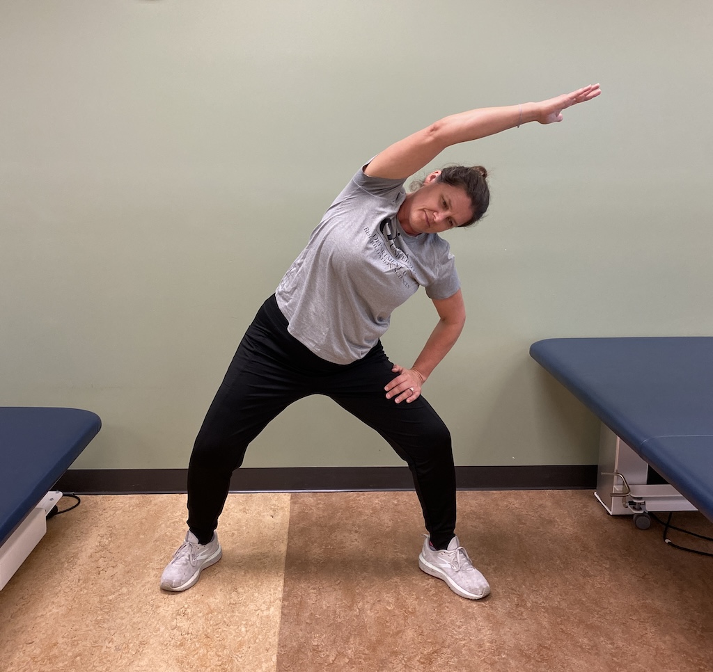 Dr. Patti Bauer, assistant professor of rehabilitation sciences and advisor for Exercise is Medicine On Campus, is wearing athletic clothing (black bottoms and a grey t-shirt) while performing a stretch. With both feet wide apart, she places her left hand on her thigh and her right hand towards the left side pointing towards the ceiling.  