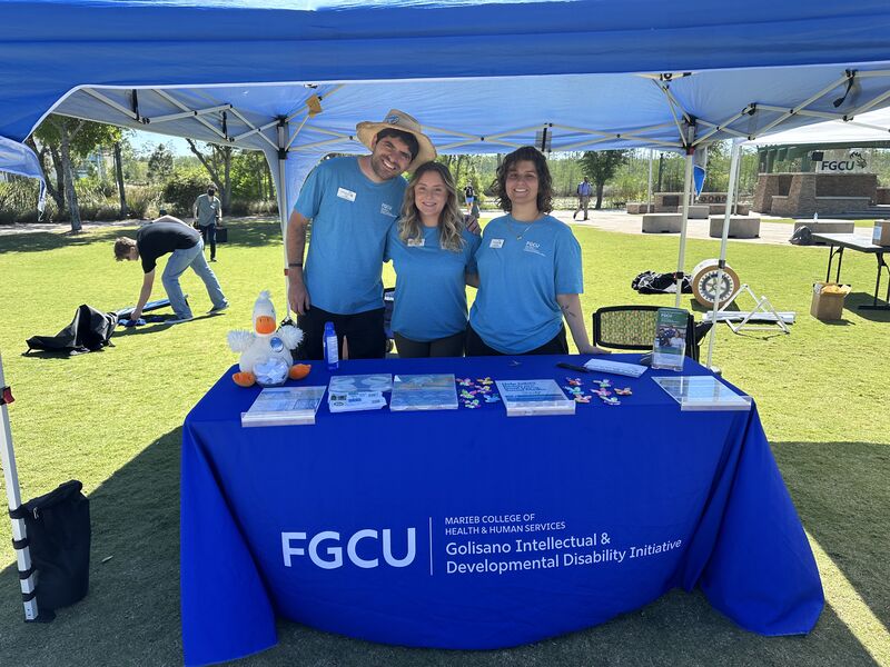 FGCU students and staff, wearing light blue t-shirts and black pants, are standing underneath a white tent at FGCU’s library lawn. They are raising money for Golisano Intellectual & Development Disability Initiatives (GIDDI) during Give Day 2025.  