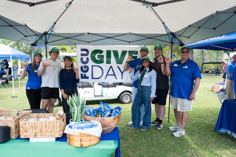 Group of FGCU students and employees wearing blue clothing stand under a tent at a table with baskets filled with snacks, in front of a sign that reads “FGCU Give Day,” outdoors on a grassy lawn.