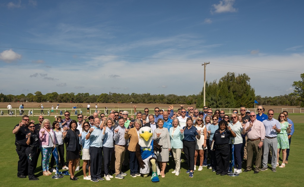 A large group of people, including FGCU employees and FGCU’s eagle mascot Azul, gather outside on a grassy area for a group photo while doing the Wings Up sign. 