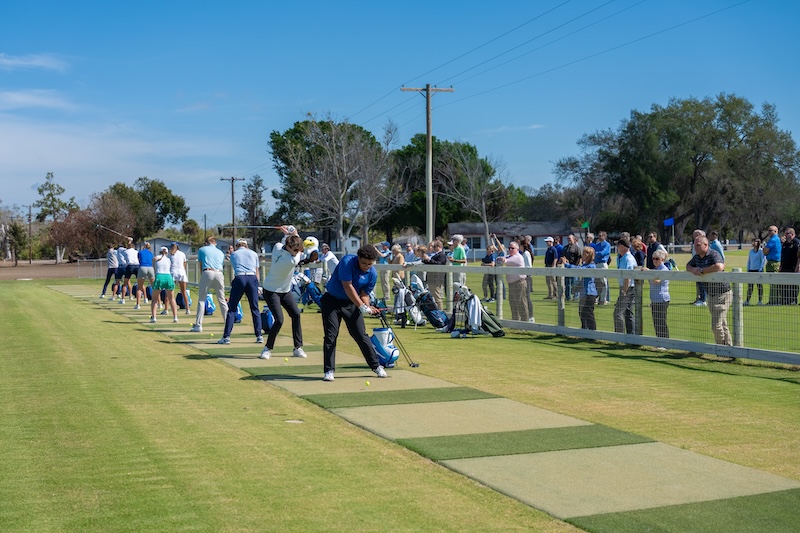  Dressed in golf attire, FGCU students are lined up on a driving range as a crowd watches from behind a white fence.