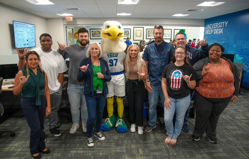 Help Desk staff members are posing for a picture with Azul for the grand opening of its new location at the Wilson G. Bradshaw Library. Azul is wearing a white jersey set with the letters FGCU and the numbers 97 in blue and outlines in green on the front of the jersey shirt.  