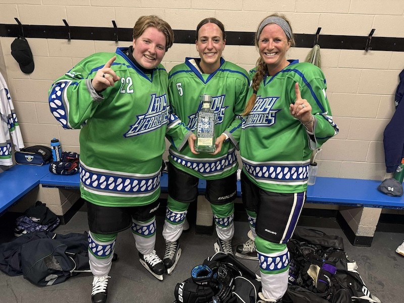 Three members of the Lady Everblades hockey team stand in a locker room wearing green jerseys. Each player holds up one finger in celebration, and the center player proudly displays a trophy.