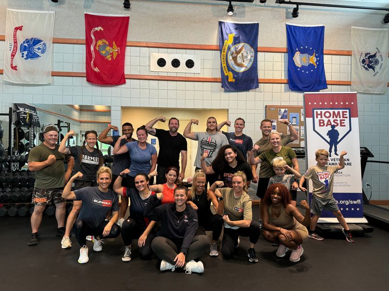 19 people wearing athletic attire are posing for a picture at a gym. Military flags can be seen in the background hanging from the ceiling. A Home Base standing banner is to the right side of the group. Gym equipment can be seen on both the right and left side of the group. 
