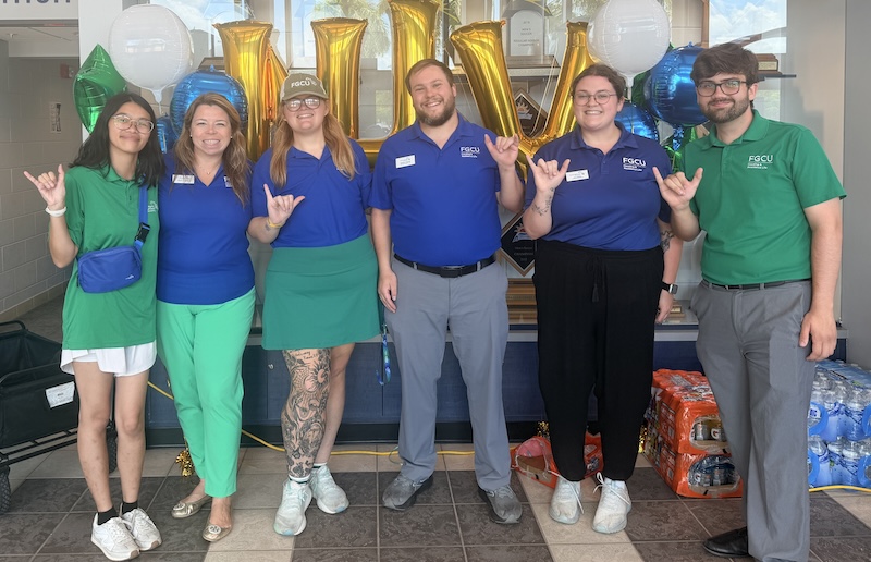 Six people wearing green and blue FGCU apparel stand in a row inside Alico Arena, posing in front of gold, blue, green and white balloons. A trophy display case is behind them, and cases of bottled water are stacked nearby.