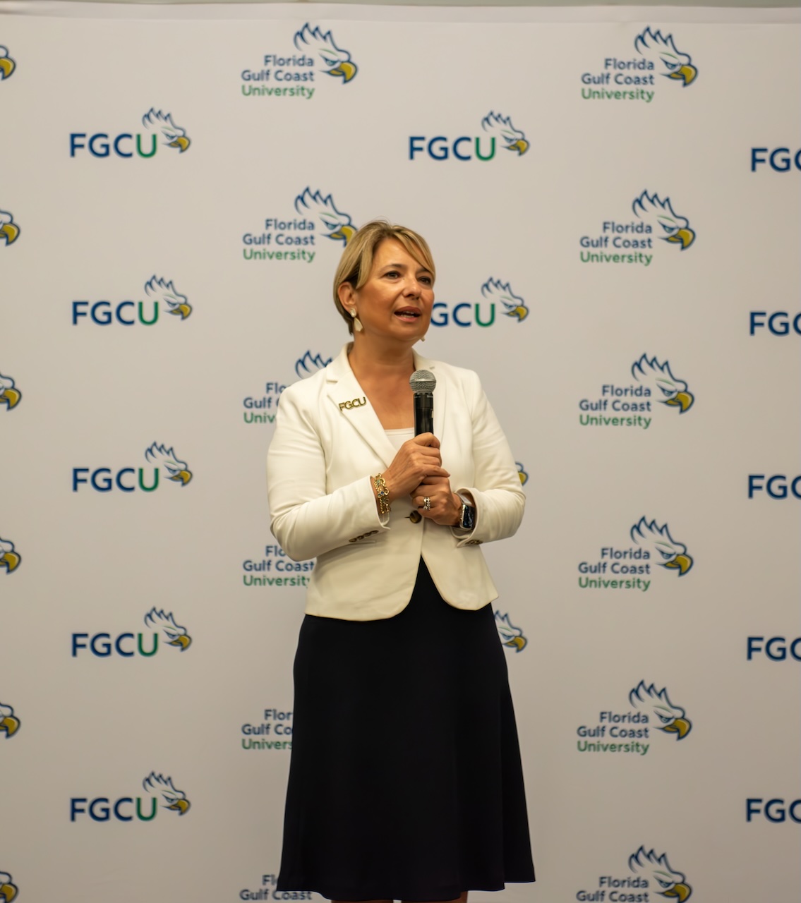 President Aysegul Timur stands in front of a Florida Gulf Coast University (FGCU) backdrop featuring repeated logos and text, including the FGCU eagle head and the words 'Florida Gulf Coast University.' She is holding a microphone and speaking at the Promotion & Grants Reception. She is wearing a white blazer with an 'FGCU' pin on the left lapel, a dark-colored dress, earrings, a bracelet and a watch. 
