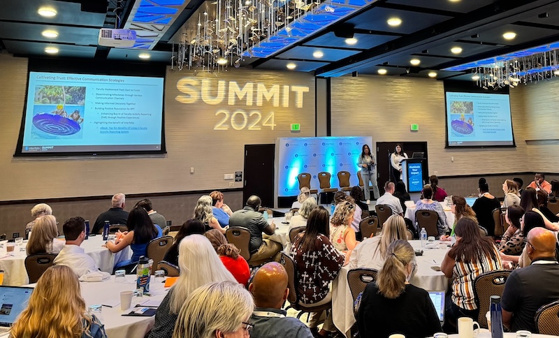 Andi Clemons, director of academic affairs, administration and budget and Kristin Toth, academic personnel coordinator are presenting to a crowd at the 2024 Interfolio Summit. Standing on a stage and in front of a podium, Andi Clemons is holding a microphone with her right hand and is wearing a light blue pant suit and white sneakers. Standing to the right of her is Kristin Toth. She is wearing a white long-sleeved blouse, black pants and white sneakers. Four empty chairs are directly behind them and a backdrop with the Interfolio logo. There are multiple round tables in front of them with people occupying them.  