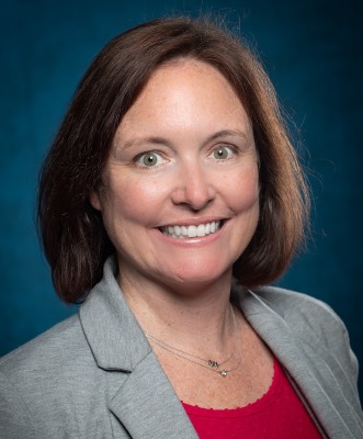 A professional portrait shows a person with short dark hair wearing a gray blazer over a red shirt. The image is framed from the shoulders up against a blue gradient background. 