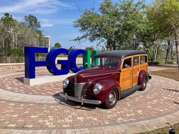A classic maroon and wood-paneled 1940 Ford Woody Wagon on a paved area at FGCU’s Library Lawn is in front of large, blue and green letters spelling "FGCU." The background includes trees and a clear blue sky.  