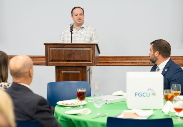 A person wearing a light-colored, short‑sleeve plaid shirt stands at a wooden podium and speaks into a microphone during a formal event. Attendees seated at round tables—set with green tablecloths, plates, glassware, and FGCU‑branded table decoration—listen from the foreground. 
