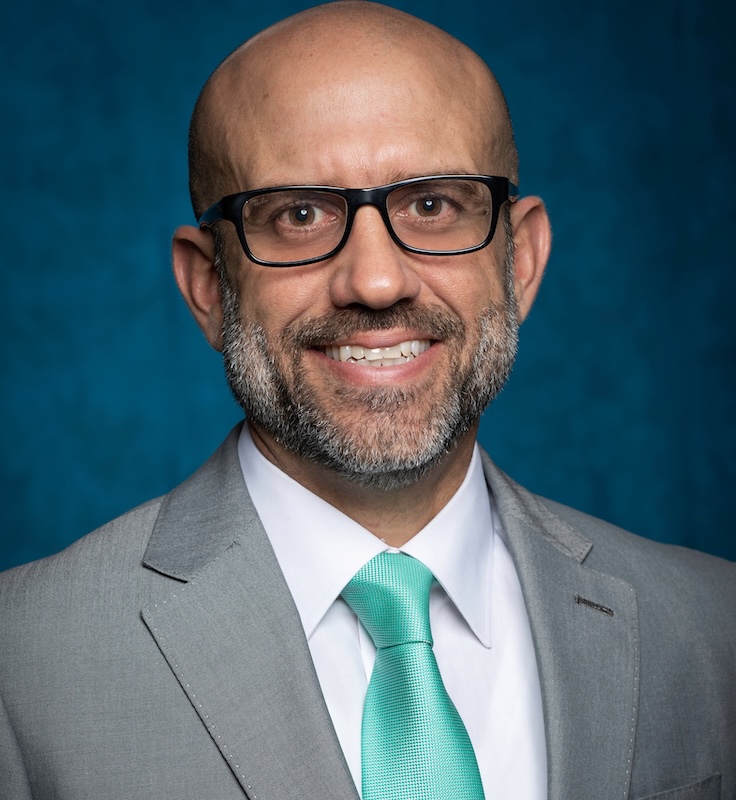Headshot of Jorge Lopez. Jorge has black glasses on, and is wearing a white shirt, a green tie, and a grey blazer. 