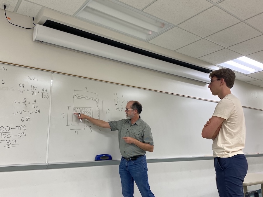 Two individuals in a classroom stand in front of a whiteboard. One person, wearing a light gray short-sleeved button-up shirt and blue jeans, points to a diagram while the other, dressed in a beige short-sleeved T-shirt and dark blue pants, observes. The whiteboard displays mathematical equations. 