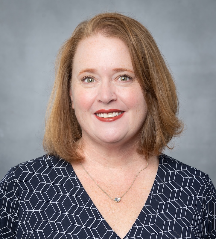 Headshot of Kristi Carman with shoulder-length hair, wearing a dark blue top featuring a white geometric pattern. She is wearing a necklace with a small pendant. The background is plain gray, providing a neutral and professional setting.