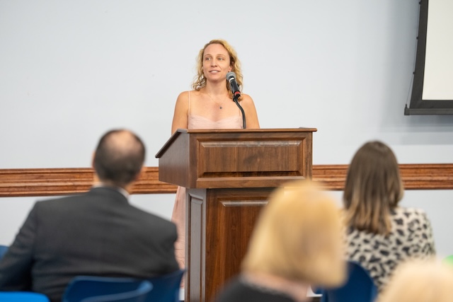 A person with blond curly hair is wearing a light pink dress and stands at a wooden podium speaking into a microphone during an indoor event. Guests seated at tables listen from the foreground. 