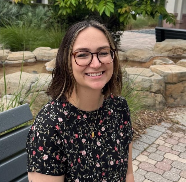Lexi Velte is wearing glasses and a black shirt with a floral pattern, sitting on a bench outdoors. The background features green plants, rock and a paved walkway, creating a natural and serene setting.