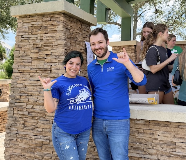 Two people wearing blue FGCU-branded shirts stand together outdoors under a pavilion, giving the “Wings Up” salute. Several attendees gather at a table in the background.