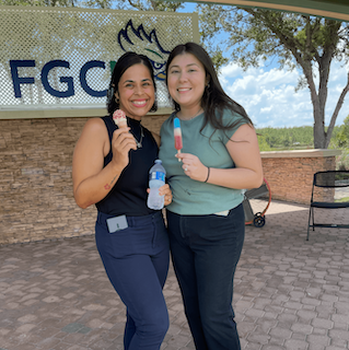 Two people pose together under a pavilion. One holds an ice cream cone and a bottle of water, while the other holds a red-and-blue popsicle. A campus sign and trees are visible in the background. 