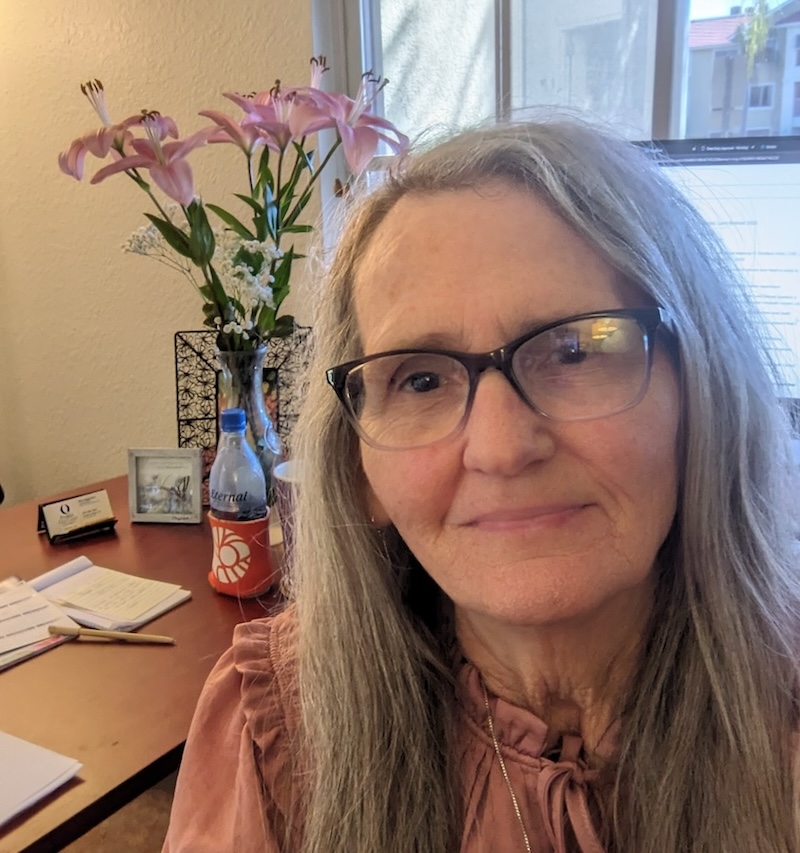 Close-up of Mary Eggleston. Mary has long pepper-colored straight hair and wears glasses and a pink blouse. Pink lilies in a vase can be seen in the background as well as a partial view of windows.