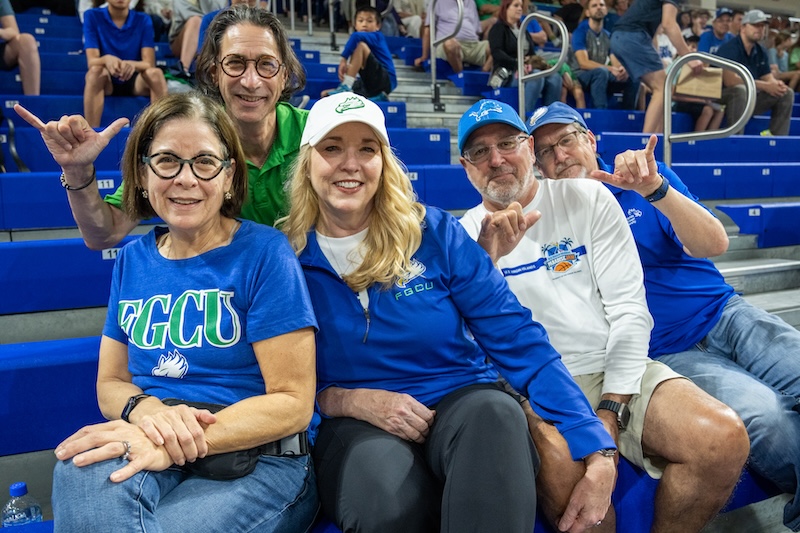 Top row, from left to right: Larry Bender is wearing a green polo shirt and giving the “wings up” salute. Bottom row, from left to right: Deb Bender is wearing a blue T-shirt with the letters FGCU in green and the FGCU eagle logo at the bottom, and jeans; Kathryn Robinson Kinsey is wearing a white cap with the FGCU eagle logo, a blue quarter-zip sweater, and black pants; Dennis Wright, is wearing a blue cap, a white long sleeve shirt and khaki shorts; Dave Anderson is wearing a blue FGCU cap, a blue FGCU polo shirt and jeans while he gives the “wings up” salute.  They are sitting on the bleachers at Alico Arena. People can also be seen in the background watching the basketball game. 
