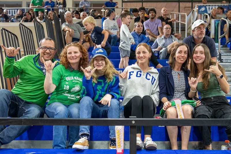 From left to right, Jeffery Rhodes, Dr. Lyndsay Rhodes, Juliette and Arabella Rhodes, Anna Carlin, and Darcy Carlin are sitting on the bleachers giving a “wing up” salute while wearing FGCU merch and green and blue clothing. People can be seen in the background sitting on the bleachers as well.  