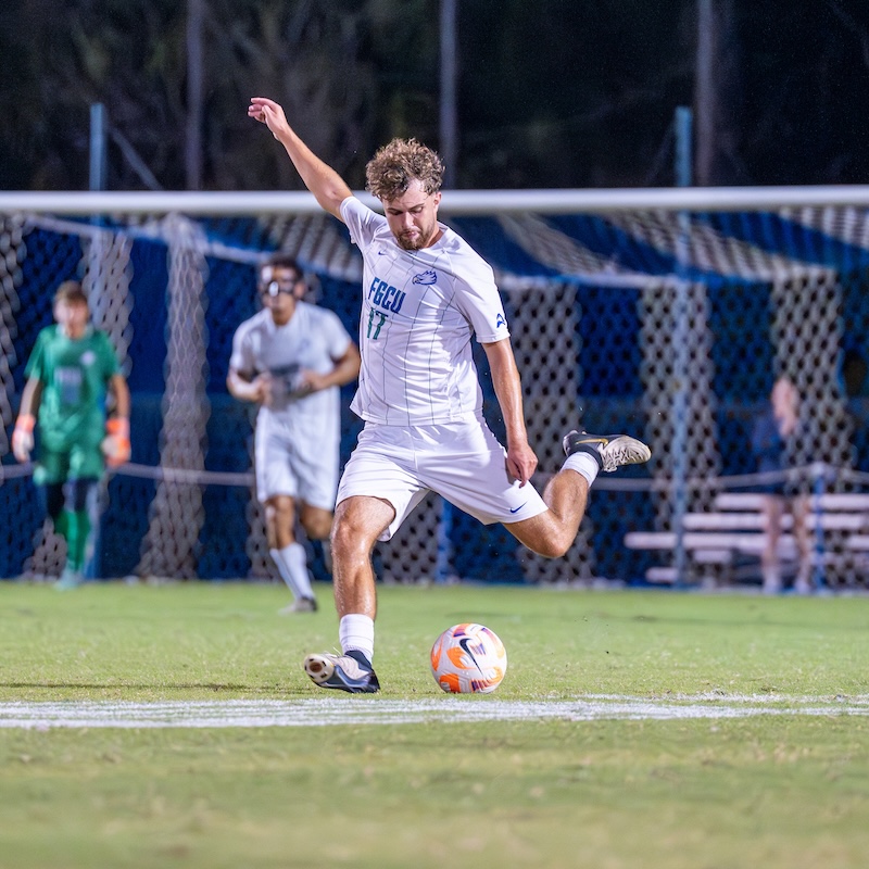 FGCU soccer player wearing a white FGCU uniform with the number 17, mid-action preparing to kick a soccer ball on a field. Other players and a goalkeeper in green are visible near the goalpost, with netting in the background.