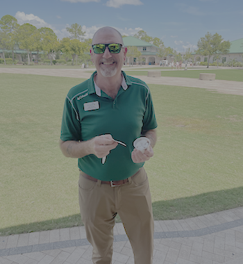 A person wearing a green collared shirt, tan pants and sunglasses is holding a dessert cup and spoon. A grassy lawn, trees, and campus buildings are visible in the background