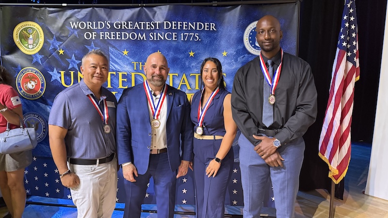 Four individuals pose together in front of a blue backdrop displaying U.S. military seals and the text “World’s Greatest Defender of Freedom Since 1775.” Each individual wears a medal, and an American flag stands to the right.   